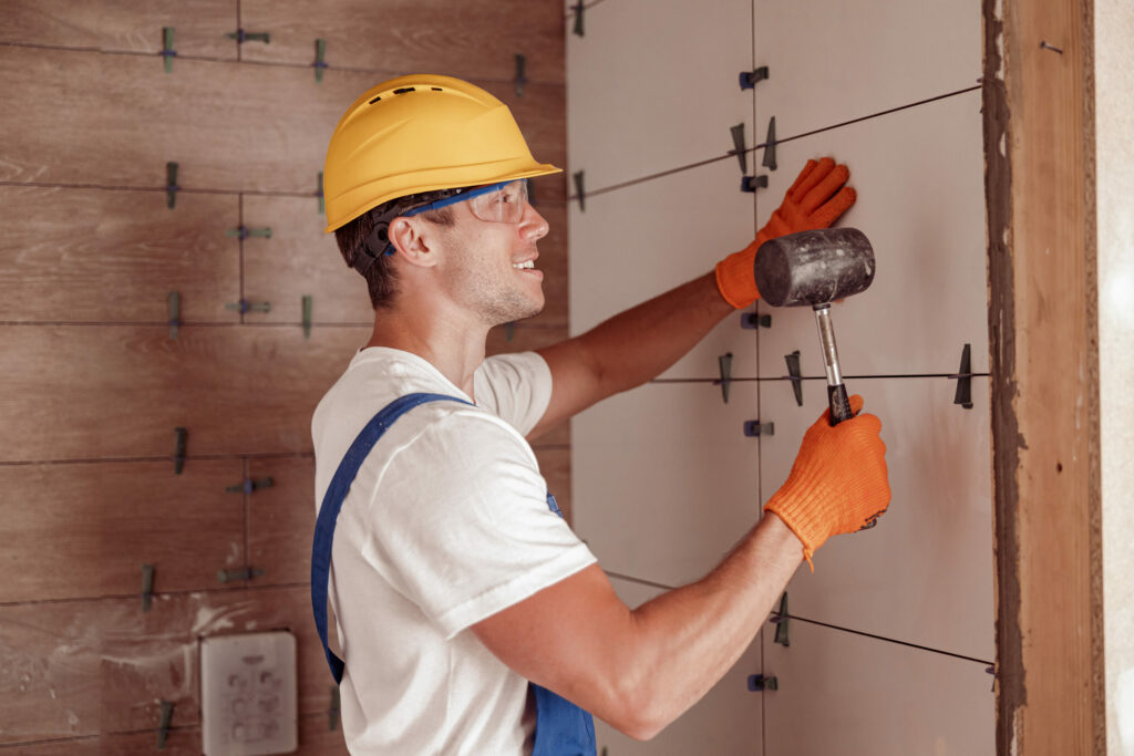 Cheerful young man construction worker using rubber hammer while placing tile on the wall